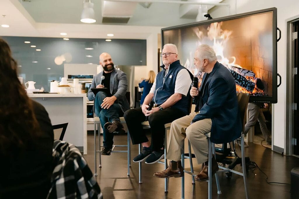 Three men are sitting on stools in front of a fireplace, engaged in conversation.